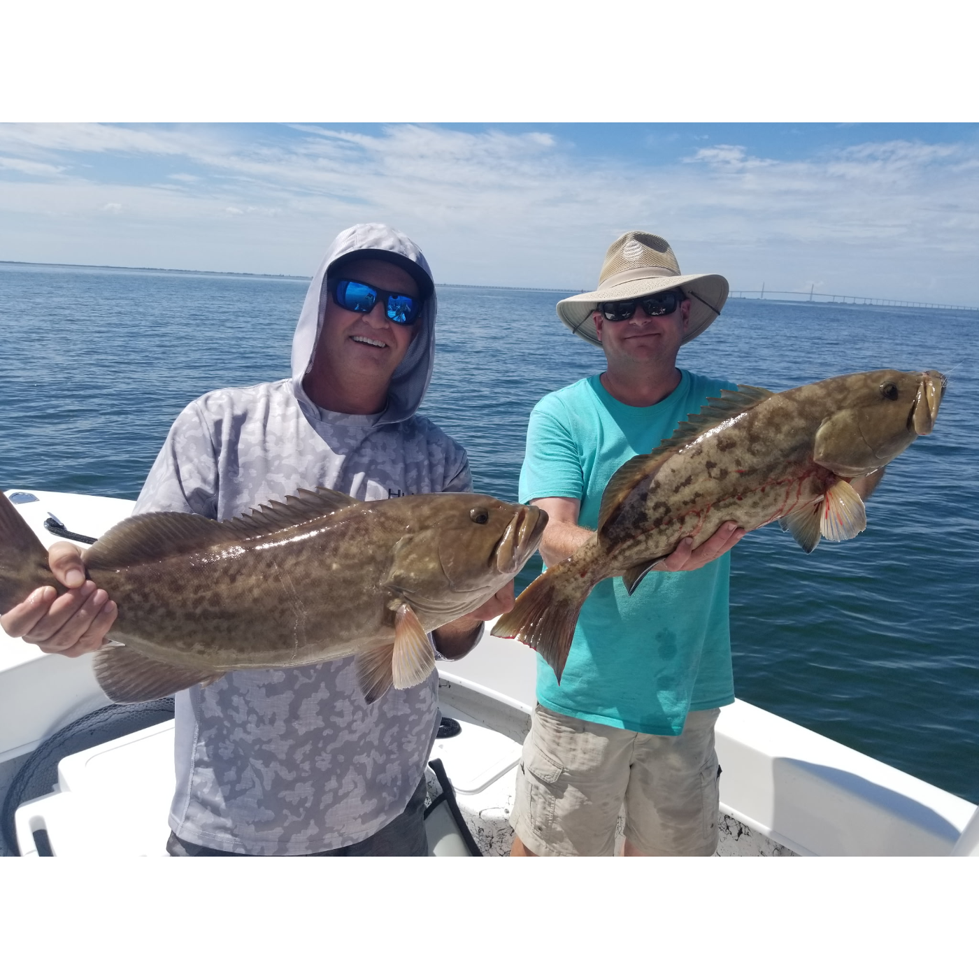 Two men on a boat holding large fish with a clear blue sky and ocean background.