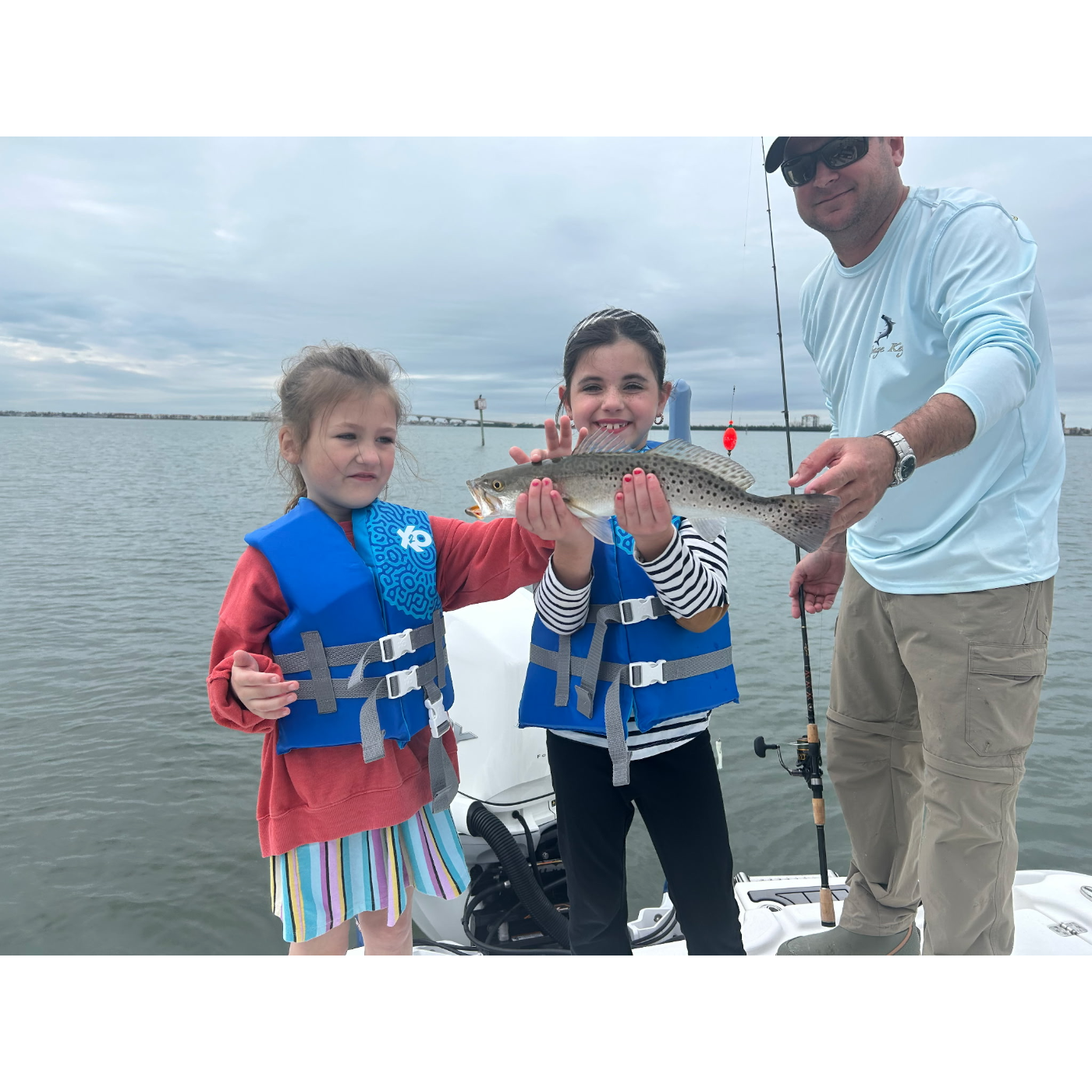 Two children in life vests holding a fish, with a man on a boat in the background.