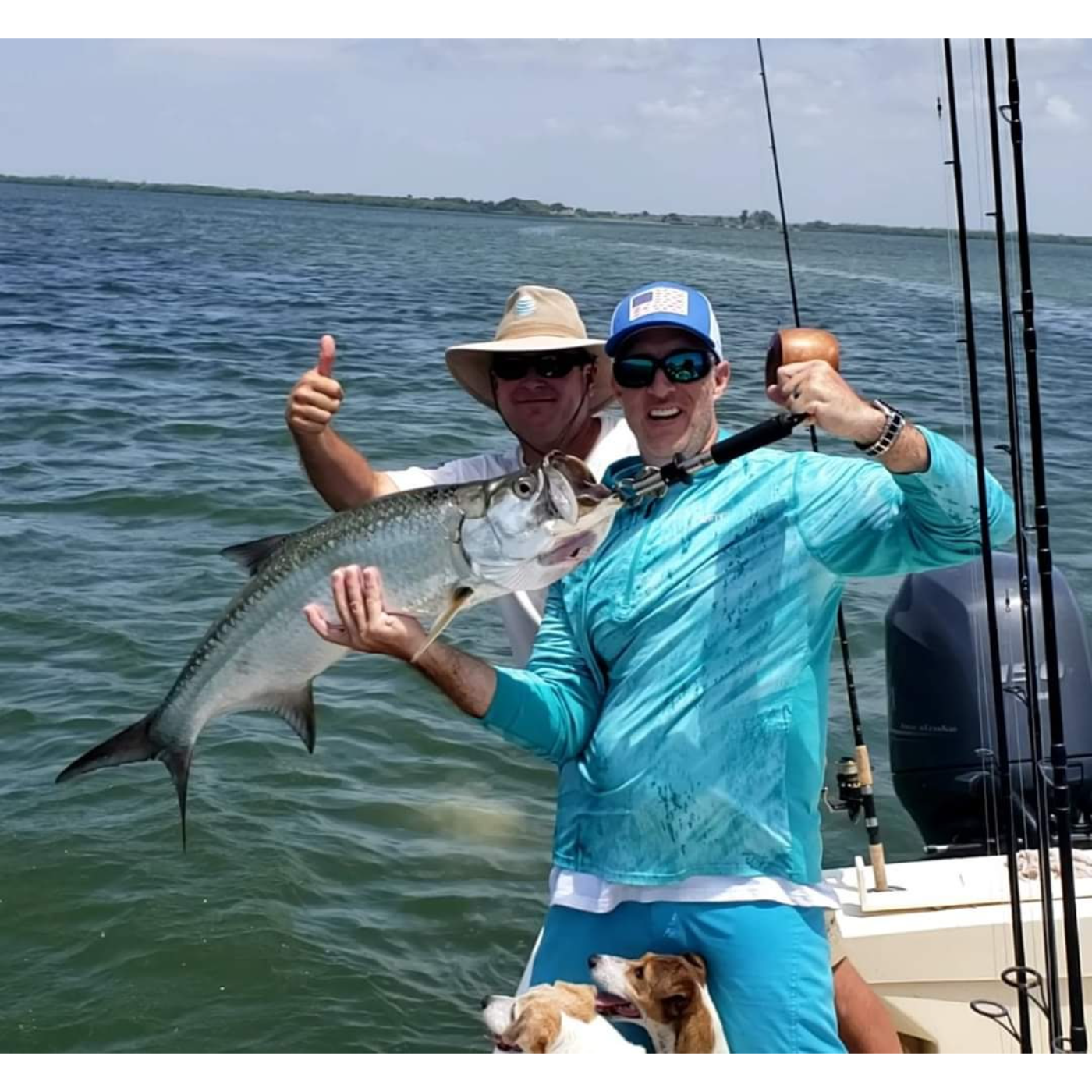 Two men on a boat with one holding a large fish, wearing sunglasses and a blue shirt, with a dog sitting nearby.