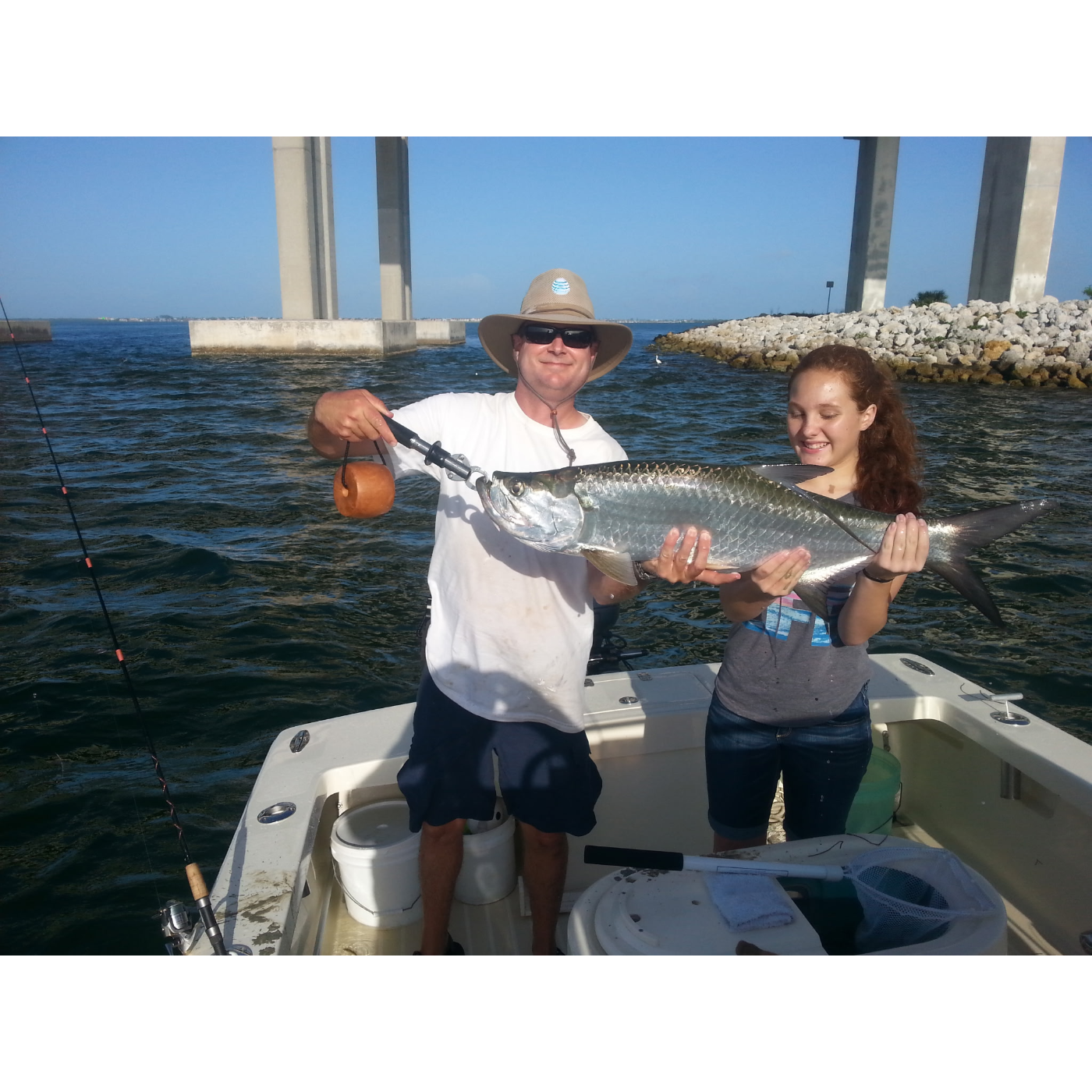 Two people on a boat holding a large fish with a clear blue sky and water background.